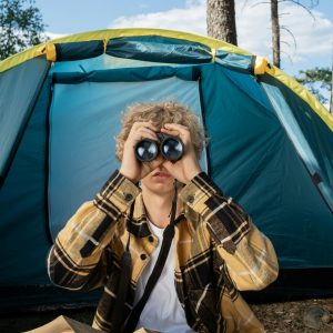 A teenager sits outside a tent holding binoculars, enjoying a summer camping trip.