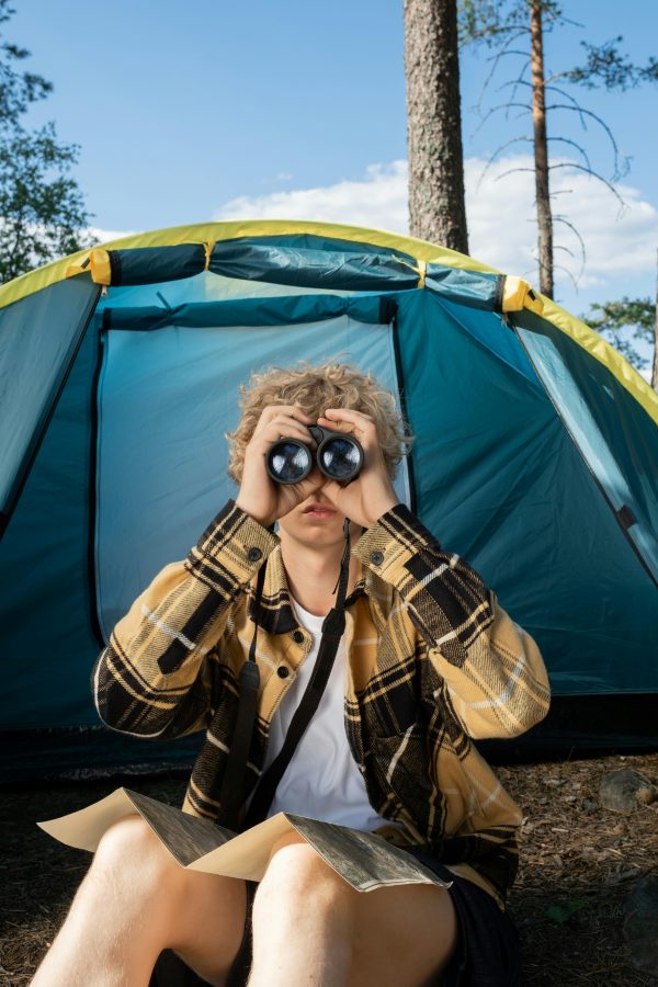 A teenager sits outside a tent holding binoculars, enjoying a summer camping trip.